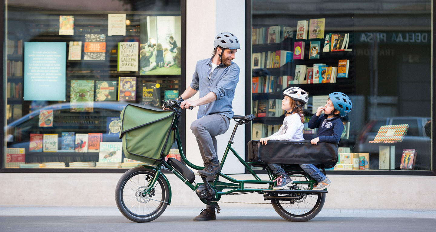 A man sitting on a green Bicicapace Justlong cargo bike with 2 children on the tail