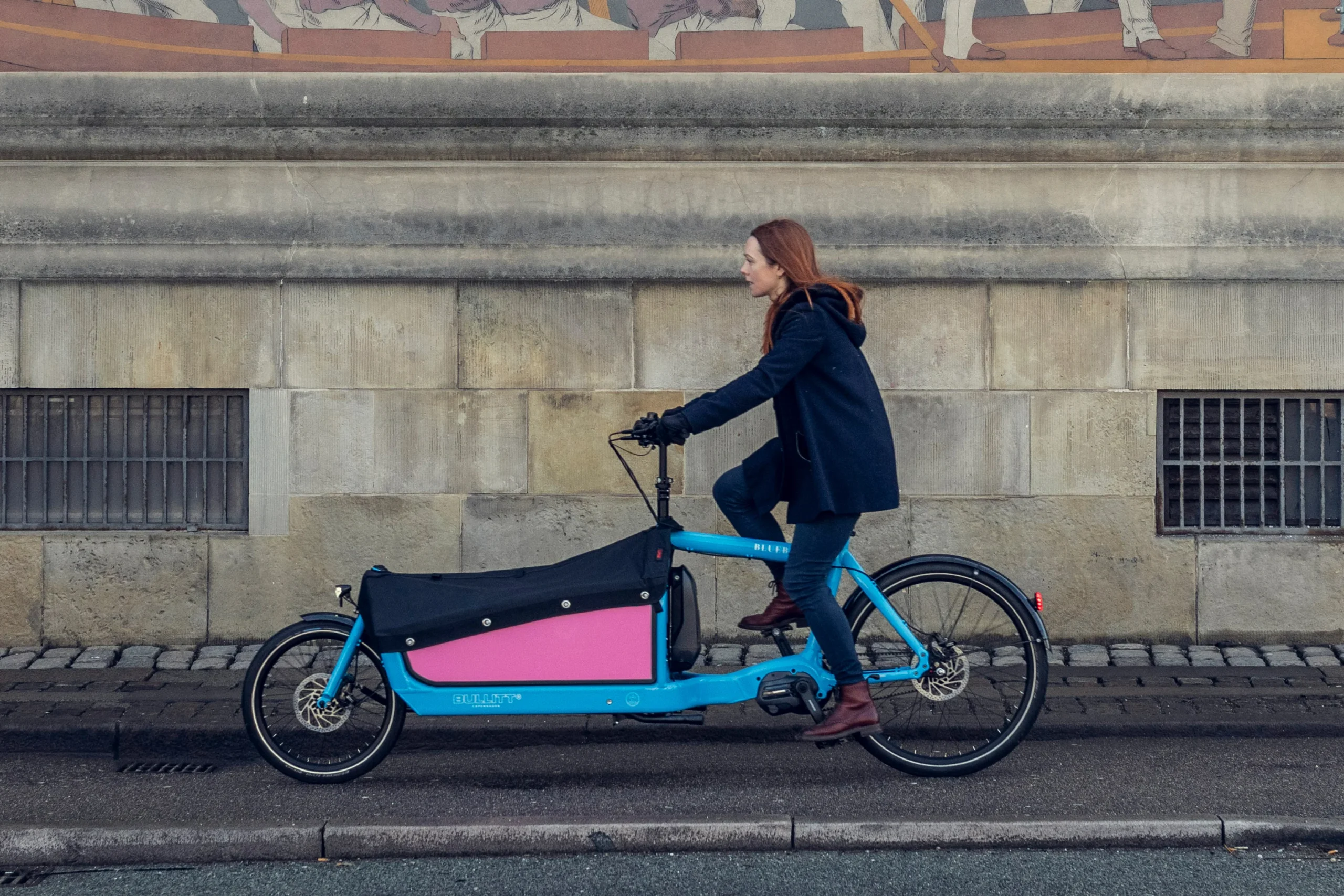A woman riding a Bluebird colored Bullit cargo bike in a urban enviroment